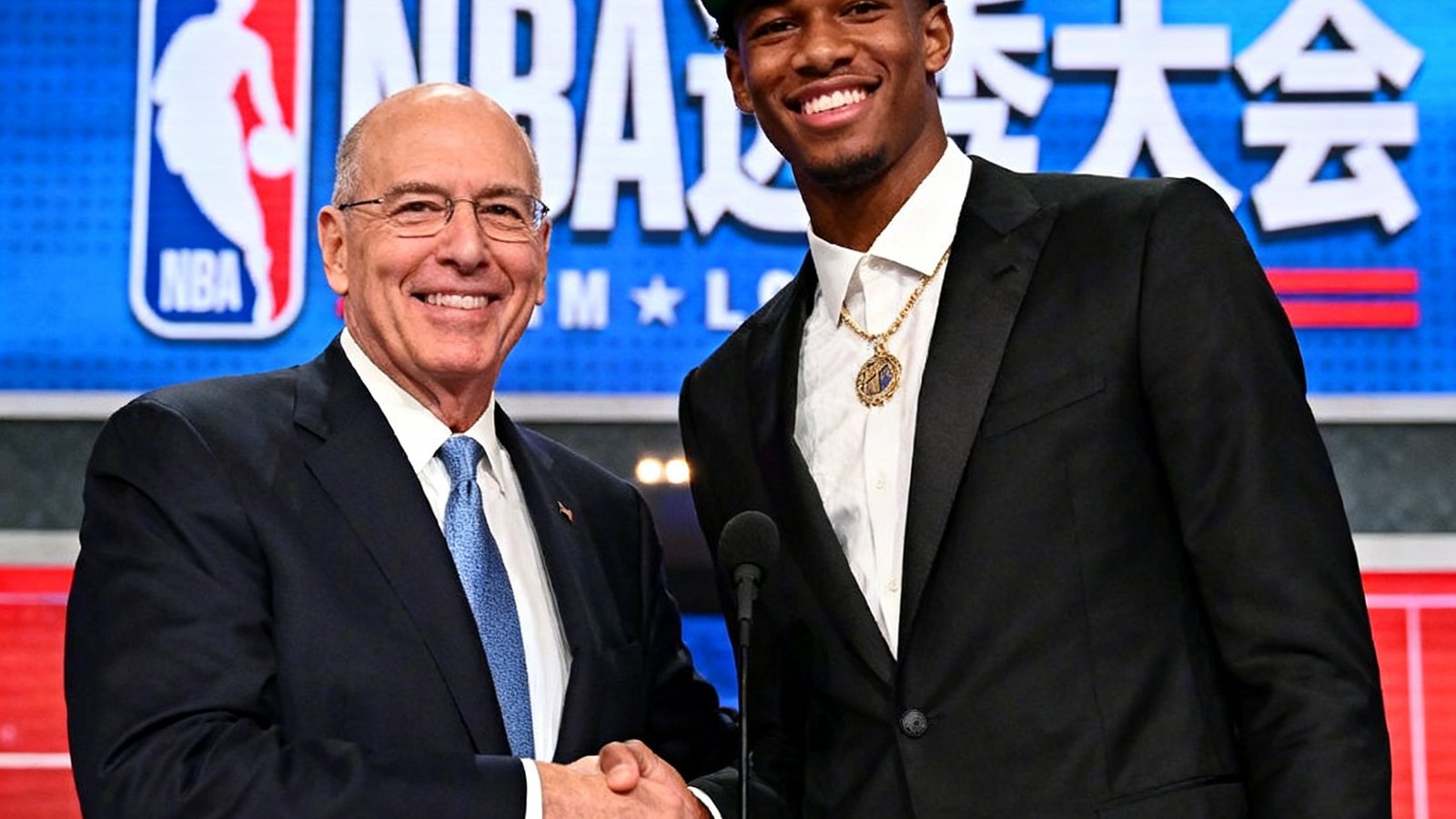 NBA选秀大会现场照片，新秀球员戴着球队帽子与NBA总裁握手合影，背景是选秀大会的标志性舞台和灯光。